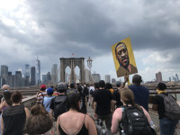 Eine junge Person spaziert mit einem Keyboard in der Hand und Kopfhörern auf dem Kopf einen Fluss entlang. Im Hintergrund ist die Skyline von New York zu erkennen.
